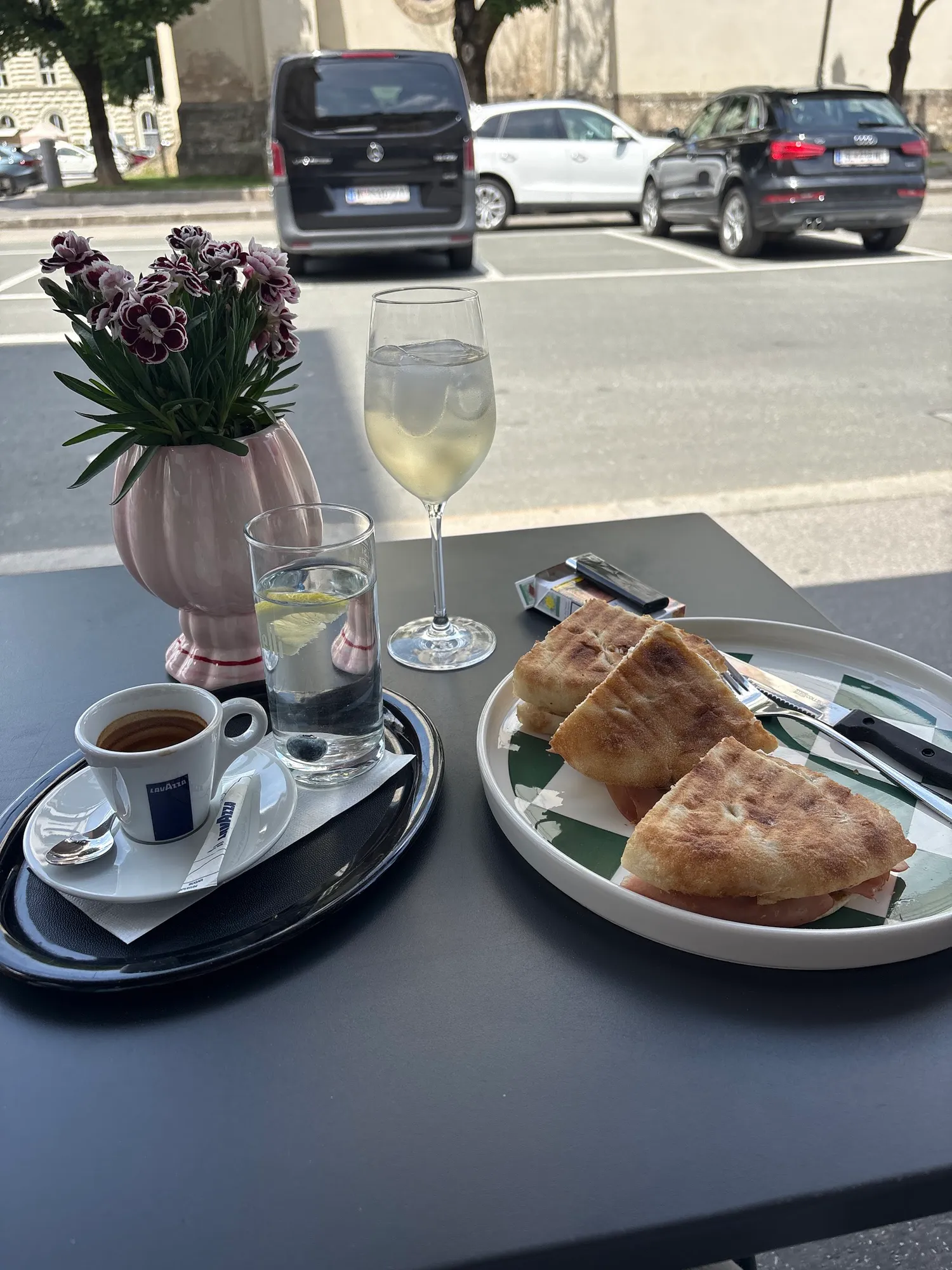 Draußen am Tisch: Espresso, Wasser mit Zitrone, ein Stielglas und Focaccia, daneben eine Vase mit Nelken