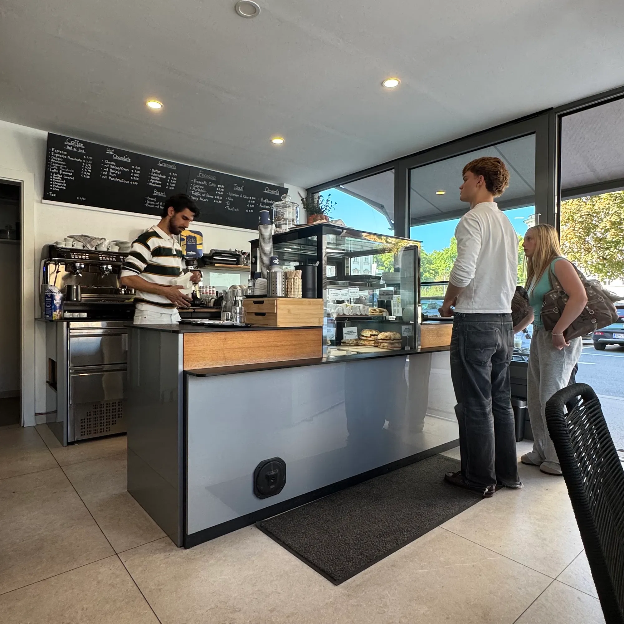Innenraum mit Theke, Kaffeemaschine, Vitrine und großer Kreidetafel, durch die Fenster der Platz