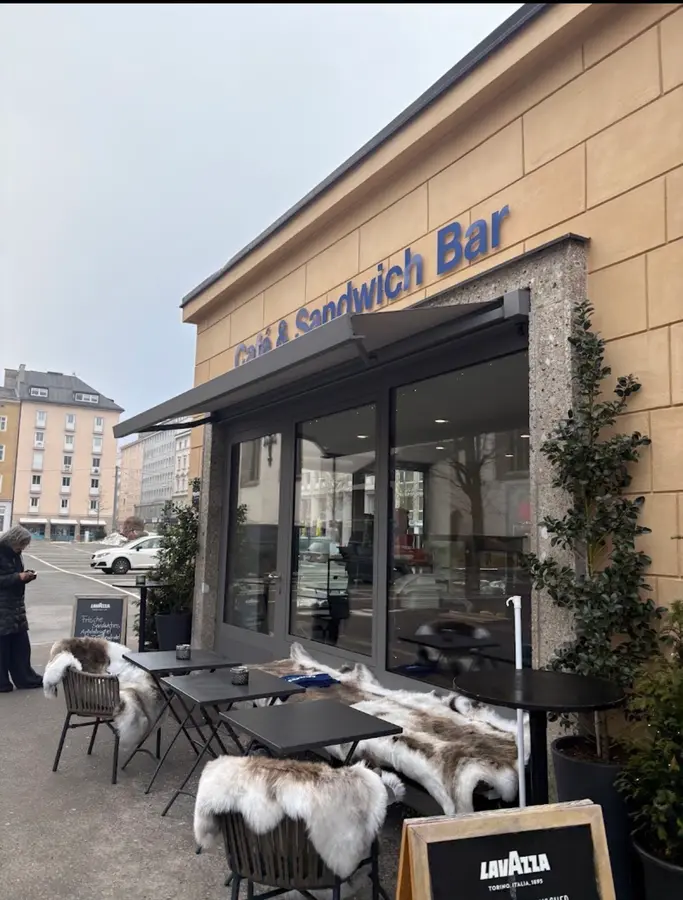 Die Glasfront mit dem blauen Schriftzug „Café & Sandwich Bar“ und Tischen mit Fellen davor